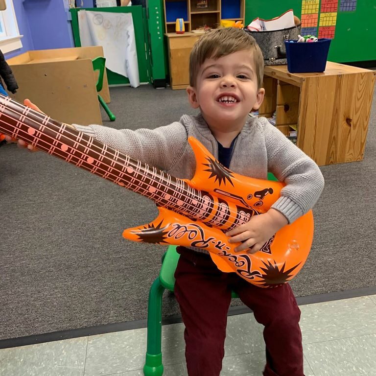 Little Toddler Boy smiling with an orange guitar and sitting in a green chair